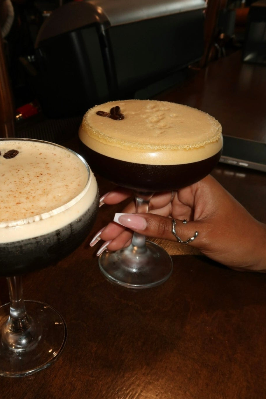 Two espresso drinks with a hand holding one on a wooden table.