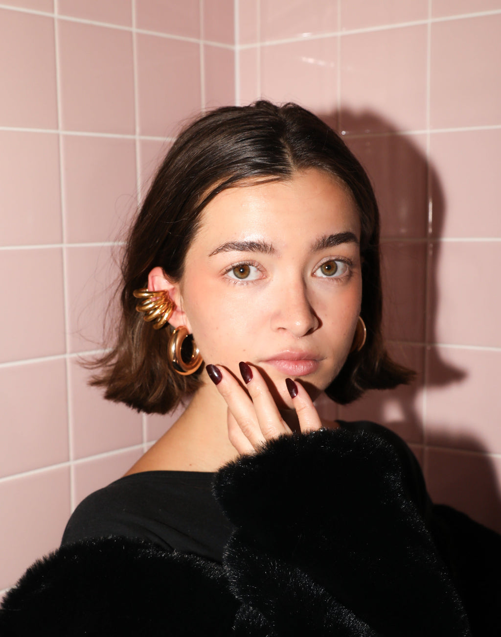 A person wearing a black furry jacket and a one-of-a-kind ear cuff with a pink tiled background.