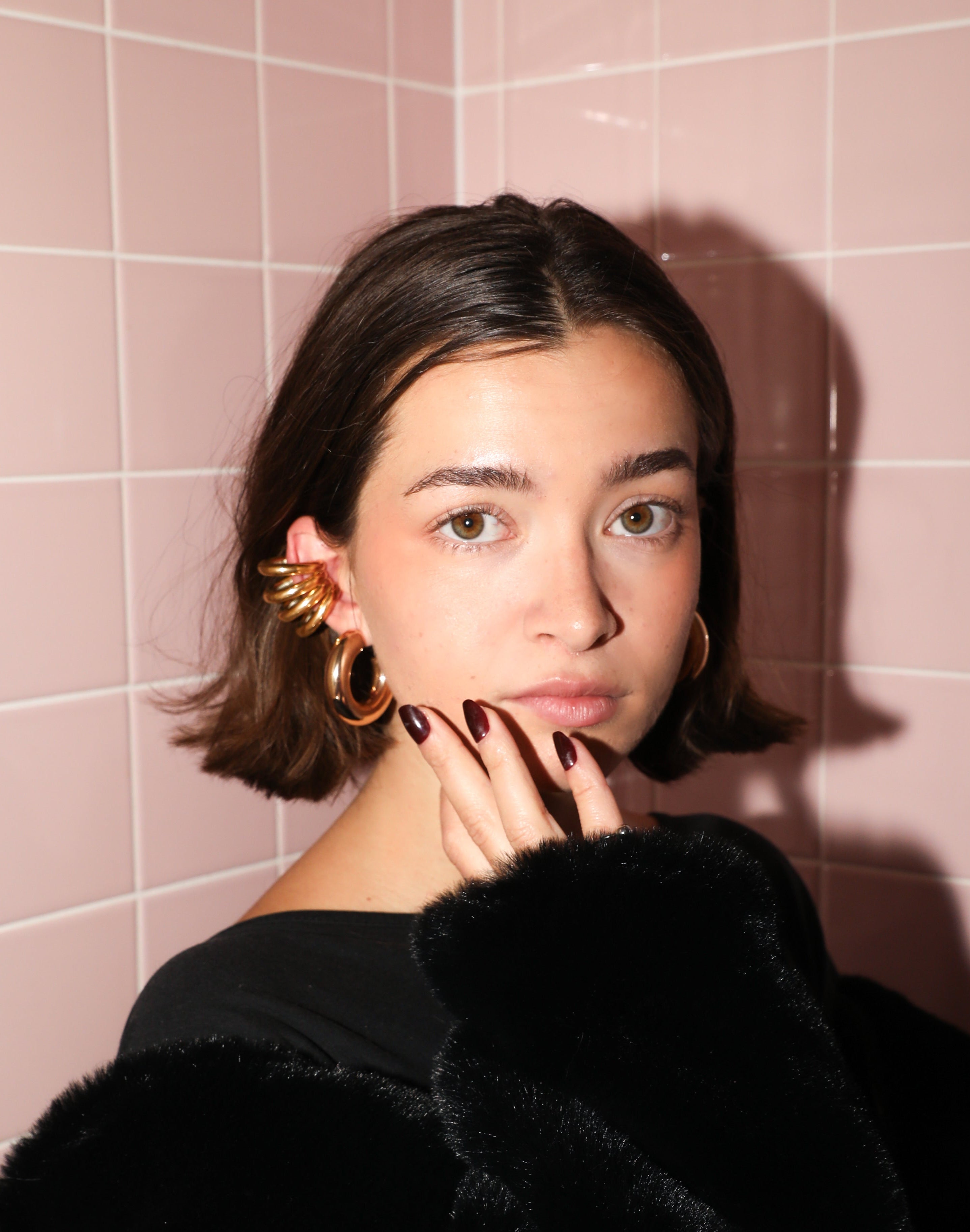 A person wearing a black furry jacket and a one-of-a-kind ear cuff with a pink tiled background.